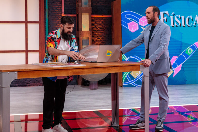 La imagen muestra a dos hombres en un set de televisión. Uno de ellos está manipulando un dispositivo sobre una mesa de madera, mientras el otro observa y escucha con atención. El hombre que está interactuando con el dispositivo lleva una colorida camisa estampada, barba y zapatillas blancas. El otro hombre, de pie a su lado, viste un traje azul claro con zapatillas deportivas y tiene una expresión seria pero relajada. En la mesa hay un portátil con un adhesivo que dice "Órbita Laika". Al fondo, una pantalla grande muestra la palabra "Física" acompañada de gráficos coloridos, como cohetes y formas geométricas, lo que sugiere que la escena está relacionada con un programa de divulgación científica.