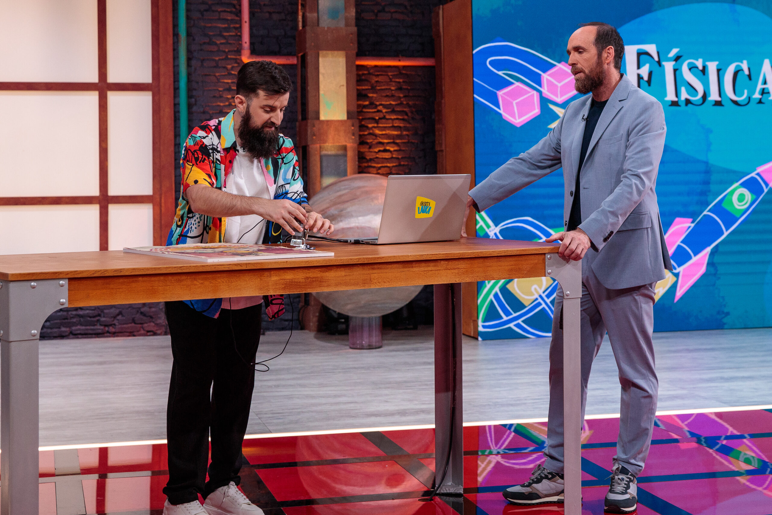 La imagen muestra a dos hombres en un set de televisión. Uno de ellos está manipulando un dispositivo sobre una mesa de madera, mientras el otro observa y escucha con atención. El hombre que está interactuando con el dispositivo lleva una colorida camisa estampada, barba y zapatillas blancas. El otro hombre, de pie a su lado, viste un traje azul claro con zapatillas deportivas y tiene una expresión seria pero relajada. En la mesa hay un portátil con un adhesivo que dice "Órbita Laika". Al fondo, una pantalla grande muestra la palabra "Física" acompañada de gráficos coloridos, como cohetes y formas geométricas, lo que sugiere que la escena está relacionada con un programa de divulgación científica.